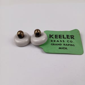 Puede incluir: Dos tiradores de armario vintage con detalles de latón y base de cerámica gris claro. Una etiqueta verde dice "Keeler Brass Co. Grand Rapids Mich."
