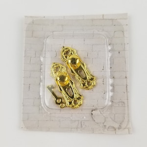 Puede incluir: Dos pomos de puerta dorados con una llave en un paquete de plástico transparente sobre un fondo de ladrillo blanco.