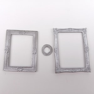 Puede incluir: Tres marcos de fotos plateados y un pequeño elemento decorativo circular se muestran sobre un fondo blanco. Los marcos tienen detalles ornamentados de estilo vintage, incluyendo patrones florales y de desplazamiento. La pieza circular tiene un diseño similar.