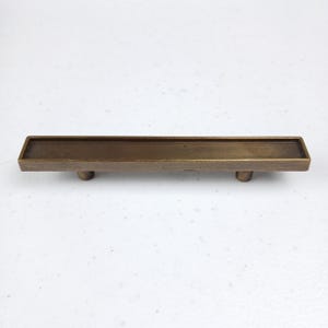 Op de afbeelding: Een rechthoekige, geborstelde messing kastgreep met een licht verhoogde rand rondom de omtrek. De greep heeft twee kleine, ronde montagegaten aan de onderkant.