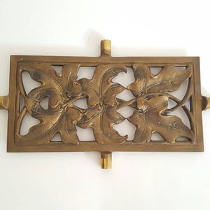 May include: A rectangular, antique brass decorative piece with an intricate leaf design. The piece has a central rectangular section with cut-out leaves and four cylindrical extensions. The overall dimensions are approximately 8 inches wide and 4 inches high.