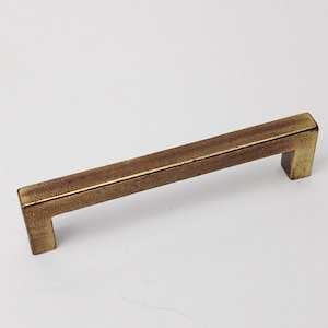 Op de afbeelding: Een rechthoekige, antiek messing kastgreep met een vierkant handvat. De hardware heeft een getextureerde afwerking en een warme, goudbruine kleur. Deze kastgreep is een decoratief element voor meubels.
