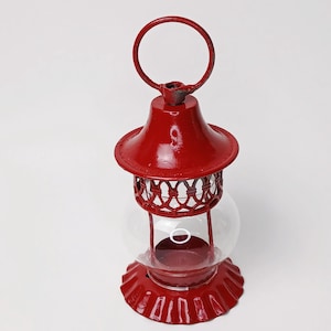 May include: A red metal lantern with a clear glass globe. The lantern features a round handle at the top and a decorative lattice design around the middle. The base is also red and has a scalloped edge. Designed to hold a candle.
