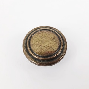 May include: A round, antique-style cabinet knob with a bronze finish. The knob features a circular design with concentric rings and a textured center. The hardware is likely made of metal and is suitable for furniture restoration or DIY projects.