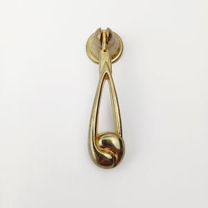 May include: A gold-toned metal zipper pull with a teardrop design. The pull features a circular top with a slot for the zipper, a slender, open-center body, and a rounded, scrolled bottom. The hardware is set against a white background.