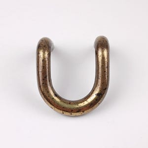 Op de afbeelding: Een U-vormige metalen handgreep met een aangetaste, verouderde messing afwerking. De handgreep heeft afgeronde uiteinden en een gebogen basis, wat suggereert dat deze voor een lade of kast kan zijn. Het metaal vertoont tekenen van slijtage.
