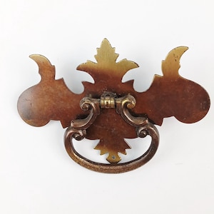 May include: Ornate antique bronze drawer pull with a decorative floral design. The pull features a curved handle and a detailed backplate with a central leaf motif and curved side elements. The bronze finish has a warm, aged patina.