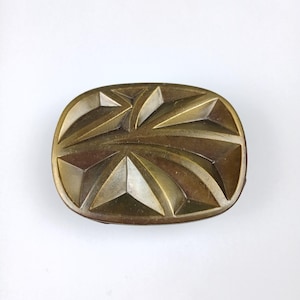 Puede incluir: Una hebilla de cinturón de metal dorado con un diseño geométrico. La hebilla tiene una superficie texturizada en relieve con un patrón de triángulos superpuestos.