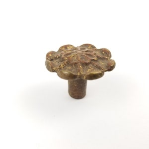Pode incluir: Uma maçaneta de armário de metal dourado com um design em forma de flor. A maçaneta tem um centro elevado com um padrão circular e um caule.