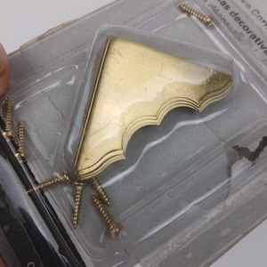 May include: A gold-colored decorative corner bracket with a scalloped edge, packaged with several gold-colored screws. The bracket is in a clear plastic container, ideal for furniture or craft projects.