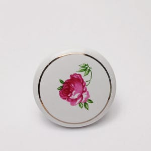 May include: A round white ceramic cabinet knob featuring a central illustration of a vibrant pink rose with green leaves. The knob is accented by a thin, circular metallic gold rim that frames the floral design, creating a polished finish.