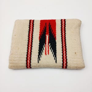May include: Cream-coloured rectangular woven cushion with a central red and black geometric design. Vertical red and black stripes flank the central design. The cushion is made of a textured fabric. Measures approximately 30 cm x 20 cm.