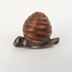 May include: A small ceramic snail figurine with a brown and white shell and a gray body.