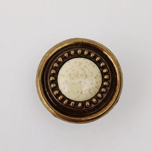 May include: A circular cabinet knob with a speckled cream-coloured centre and a brass-coloured metal rim. The rim has a series of small, raised dots around the centre. Suitable for drawers and cabinets.