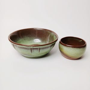 May include: Two ceramic bowls. The larger bowl has a wide rim and a light green exterior with a brown interior. The smaller bowl is a similar design, with a light green exterior and a brown interior. Both bowls have a glossy finish.