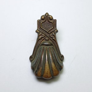 Könnte beinhalten: Ein vertikaler Möbelgriff aus bronzefarbenem Metall mit einem strukturierten, schildförmigen Oberteil und einem Unterteil in Form einer gewellten Muschel. Der Beschlag weist eine verwitterte Patina mit dunklen Vertiefungen und hellen Kanten auf.