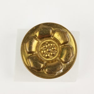 May include: A gold-coloured metal button with a raised design of a flower with a centre of small circles.
