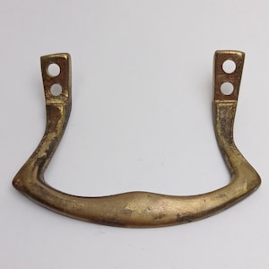 May include: A U-shaped brass drawer pull with a weathered, tarnished finish. The handle features two vertical mounting brackets, each containing two circular screw holes for attachment. The metal surface shows signs of age and oxidation.