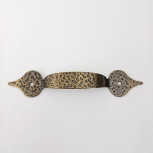 May include: A decorative bronze-colored door pull with a textured surface. The handle has a central bar with a series of small, irregular indentations, and each end features a stylized, leaf-like shape with a circular hole.