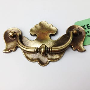May include: Antique brass drawer pull with a curved handle and decorative backplate. The pull features a central cloud-like design and ornate details, perfect for vintage furniture restoration. The overall dimensions are approximately 6 inches wide.