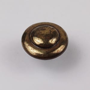 May include: A close-up of a vintage brass button with a circular design. The button has a raised centre and a worn, antique finish, showing signs of age and use. The button is a warm, golden-brown colour.