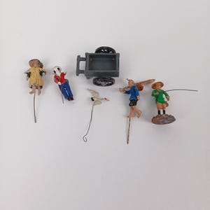 Puede incluir: Una colección de figuras en miniatura vintage y un pequeño carro. Las figuras representan personas con ropa tradicional, un pájaro y un carro. Las figuras están pintadas en varios colores, incluyendo amarillo, azul, rojo y verde. El carro es gris.