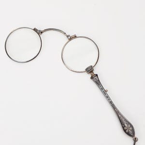 Op de afbeelding: Antieke zilverkleurige bril met ronde glazen en een decoratief handvat. Het handvat heeft ingewikkelde geëtste patronen en een kleine lus. De bril is ontworpen om aan het handvat te worden vastgehouden.