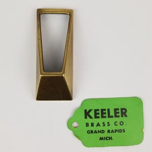 May include: A gold-toned metal door pull with a rectangular shape and a cut-out design. The pull has a tag attached to it that reads "Keeler Brass Co. Grand Rapids Mich."