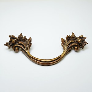 Op de afbeelding: Een gebogen metalen ladehandgreep met een antieke messing afwerking. De handgreep is voorzien van gedetailleerd, verhoogd blad- en bloemmotief aan beide uiteinden, met een glad, gebogen middengedeelte voor bevestiging aan meubilair.