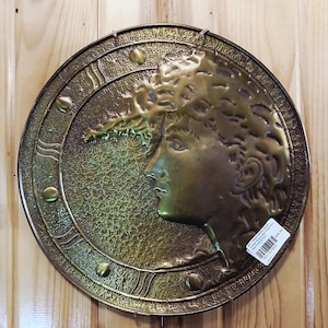 May include: A round, antique-style metal wall hanging with a profile of a woman's face. The piece is primarily bronze and gold in color, with a textured background and decorative border. The woman's hair is intricately detailed.