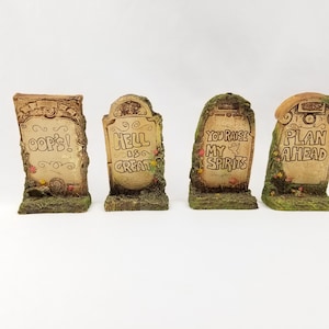 Puede incluir: Cuatro figuritas decorativas con forma de lápida con dichos humorísticos. Las lápidas están hechas de un material marrón claro y tienen una superficie texturizada. Los dichos en las lápidas son "¡Ups!", "El infierno es genial", "Tú elevas mi espíritu" y "Planifica con anticipación".