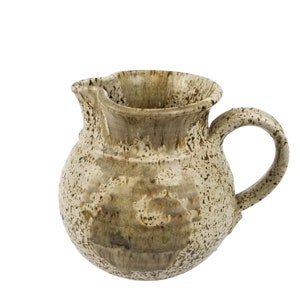 May include: A speckled brown ceramic jug with a wide, rounded body and a flared lip. The jug has a handle that curves up and over the lip.