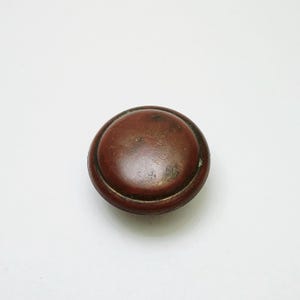 May include: A round, dark reddish-brown cabinet knob with a smooth, slightly weathered surface. The knob features a raised circular center section surrounded by a recessed border, showing signs of wear and minor surface imperfections.