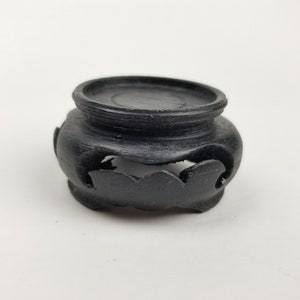 May include: A small, round, black wooden display stand with a carved, wave-like design around the base. The stand has a flat top and is designed to hold a small object. The stand is made of dark wood.