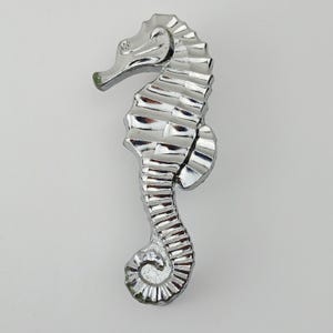 May include: A chrome seahorse ornament. The seahorse is shown vertically, with a detailed, textured surface. The head is slightly tilted upwards, and the tail is curled. The figurine is a decorative item, likely for a coastal or nautical theme.
