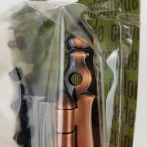 Puede incluir: Primer plano de una bisagra de color cobre con un remate decorativo. La bisagra está empaquetada en una bolsa de plástico transparente con fondo verde.