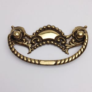 May include: A brass-toned metal drawer pull featuring a curved, rope-textured handle attached to a decorative backplate. The backplate displays intricate scrollwork, a central smooth arch, and a beaded border design with two mounting points.