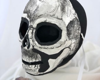 May include: A black and white skull mask with a painted bone structure. The mask is made of paper mache and has a textured surface. The mask is on a white background.