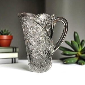 May include: Clear glass pitcher with a decorative, cut-glass pattern. The pitcher has a handle and a scalloped rim. The design features starburst and diamond shapes. The pitcher is likely used for serving beverages.