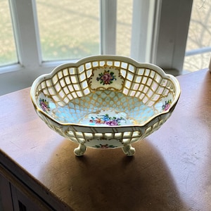 Könnte beinhalten: Eine cremefarbene Porzellanschale mit offenem Gitterwerk und gewelltem Rand. Die Schale hat einen hellblauen Innenraum mit floralen und goldenen Akzenten. Sie steht auf drei kleinen Füßen und ist wahrscheinlich dekorativ.