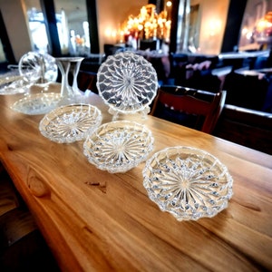Könnte beinhalten: Eine Sammlung klarer, geschliffener Glasservierschalen und -teller, die auf einem Holztisch angeordnet sind. Die Schalen haben ein Sternmuster und werden in verschiedenen Größen präsentiert. Im Hintergrund sind auch eine hohe Vase und ein Kronleuchter zu sehen.