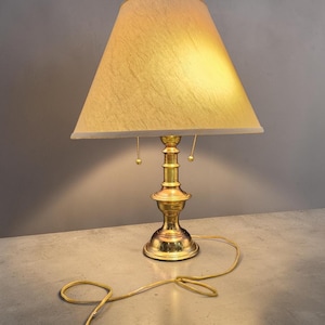 May include: A table lamp featuring a polished brass base with a tiered, pedestal-style column and a wide, circular foot. The lamp is topped with a conical, textured beige fabric shade and includes two pull-chain switches hanging from the socket.