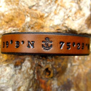 May include: Brown leather bracelet with black stitching and an anchor symbol. The bracelet is engraved with the coordinates "39° 3' N 75° 23' W".