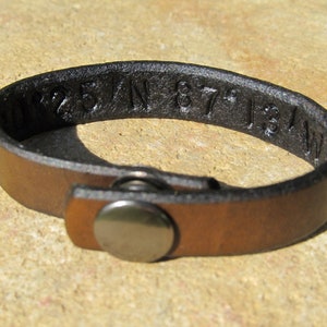 May include: A brown leather bracelet with a black leather strap and a silver metal clasp. The bracelet has the coordinates "N 25° W 87° 13'" engraved on it.