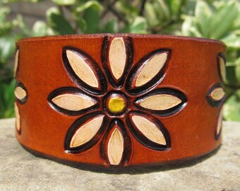 May include: A brown leather bracelet with a carved daisy design. The daisy has a yellow center and white petals.