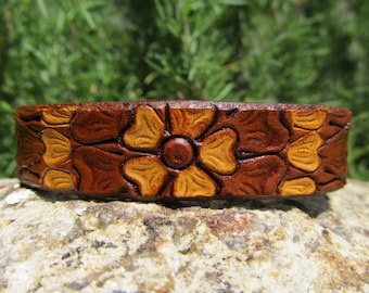 May include: A brown leather bracelet with a carved floral design. The flowers are a light brown color and have a raised center.