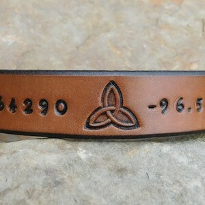 May include: Brown leather bracelet with a Celtic knot design and the coordinates 32.864290 -96.54130 engraved on it.