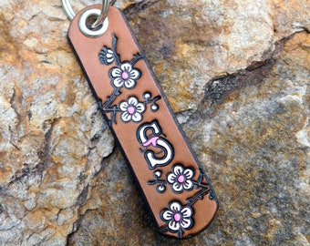 May include: Brown leather keychain with a silver key ring and a floral design. The keychain has the letter "S" in the center and pink and white flowers on a vine.
