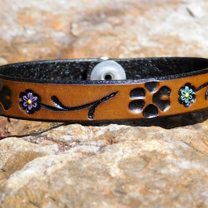May include: A brown leather bracelet with black paw prints and floral accents. The bracelet features a black interior and a silver snap closure. The flowers are painted in purple, blue, and yellow. The bracelet is approximately 1 inch wide.