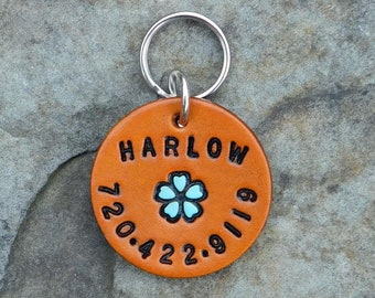May include: A round brown leather dog tag with a silver key ring. The tag is engraved with the name "HARLOW" and the date "7.20.4.22.9.11.9". A blue flower is in the center of the tag.